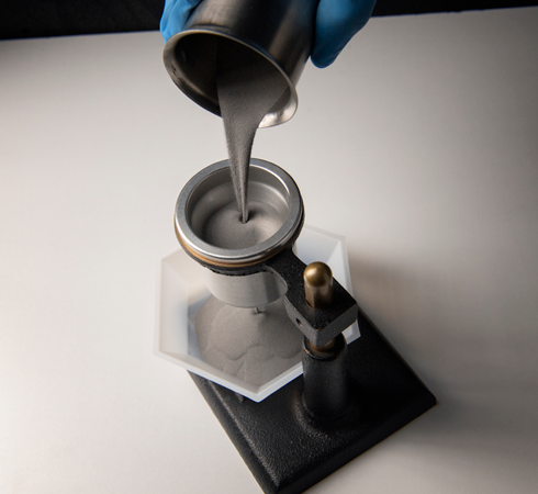 Gloved hand pouring grey liquid from metal dispenser into circular mould on black stand