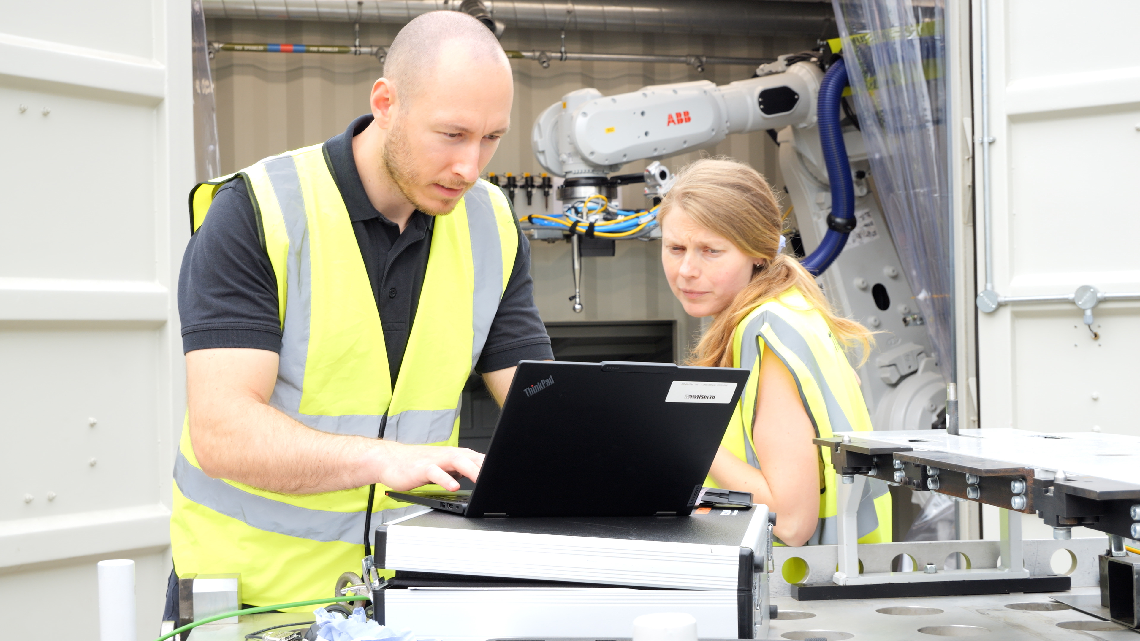Two people operating a PC in front of a shipping container with a ABB robot